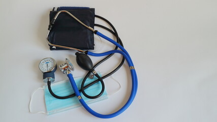 Metal phonendoscope with blue rubber tubes and a tonometer with a medical mask on a light background.