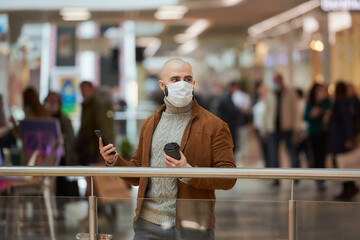 Fototapeta na wymiar A man with a beard in a face mask is looking to the right and holding a smartphone and a cup of coffee in the shopping center. A bald guy in a surgical mask is keeping social distance.