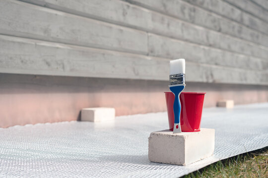 Paint Brush And A Bucket With Paint Next To A Building Exterior Wall. Painting House Plinth Outside. Home Renovation. Nobody