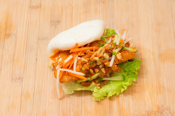 Delicious steamed bao bun on wooden table with fried chicken and fresh vegetables - salad, carrots and roasted peanuts. Popular street food