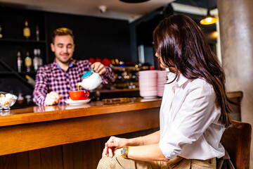 Caucasian handsome male bartender making cup of tea to brunette indian woman customer at cafe counter