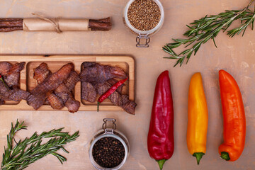Jerky snacks on wooden board, red, orange and yellow papper, meat sticks, peppercorns in glass jars and rosemary on light brown background. Top view. Dried spiced meat for beer.