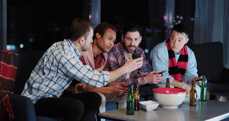 Multi-ethnic happy guys cheerinhg for football soccer team watching TV game match late at night together spending evening. Football fans. Entertainment.