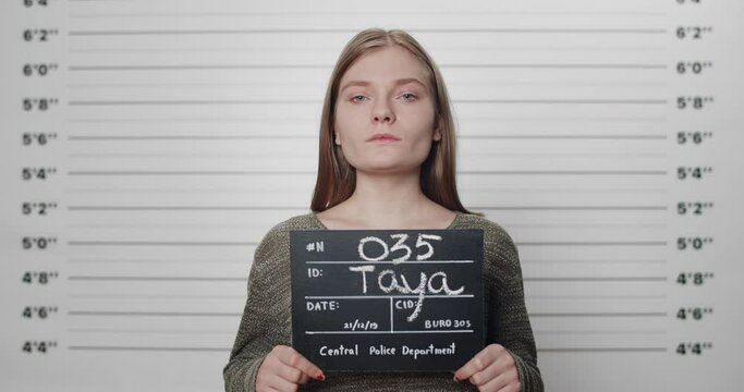 Mugshot of millennial arrested woman holding sign while being photographed in police department. Female criminal with blue eyes raising head and looking to camera in front of metric lineup wall.