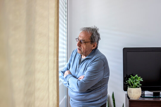 Depressed lonely senior man standing alone and looking through the window. Unhappy mature man with eyeglasses looks sadly outside the window at home. People and depression concept. Copy space.