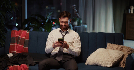 Obraz premium Happy adult young business person in suit sitting on sofa relaxing at home using application social network on smartphone screen networking online.
