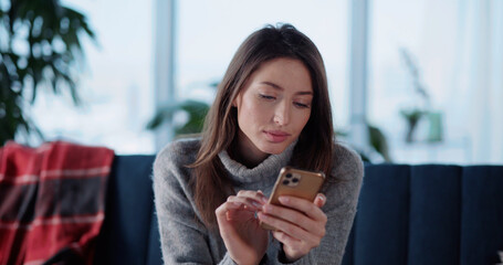 Young caucasian millennial woman using mobile phone typing cellphone messages on social media connecting to friends staying home in cozy living room.