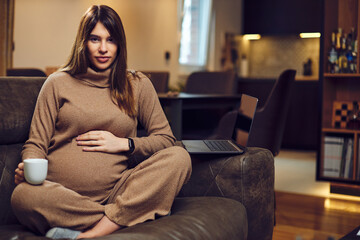 Obraz premium Cute pregnant woman sitting on sofa at home and enjoying her morning coffee caffeine-free.