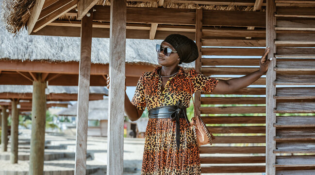 African woman wearing African suit and sunglasses standing inside a bungalow and looking out towards the beach in Ghana West Africa