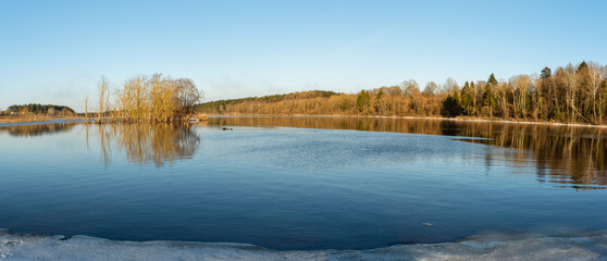 Panoramic view of the blue river. The near shore is covered with drifts of ice, the far shore is with trees and bushes without foliage. Spring evening with clear skies and sunshine.