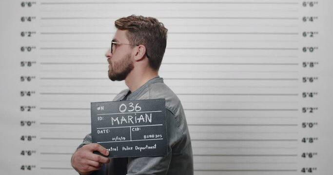Side profile mugshot of man with beard and mustache standing at metric line up wall. Male arrested in glasses turning head to camera while being photographed in police department.
