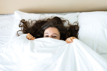 Playful young brunette woman hiding face under blanket while lying in cozy bed after waking up, looking at camera, feeling shy peeking from duvet, covering with white sheet. Good morning. Top view