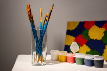 Glass with drawing brushes on table with paint banks on painting background