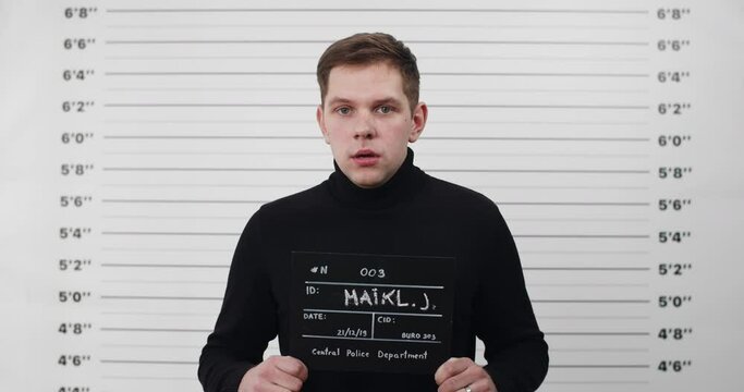 Portrait of young man in black turtleneck holding sign for photo in police department. Crop view of guy breathing deeply and posing while looking to camera. Concept of crime.