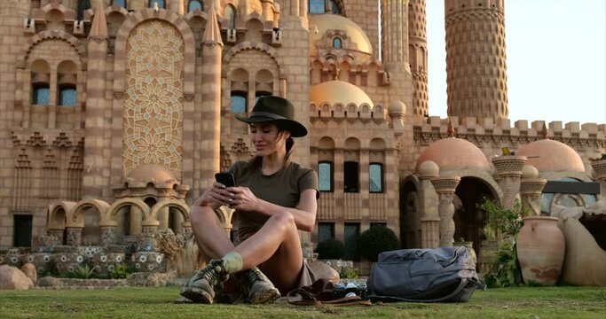 pretty tourist, in tourist boots and a cowboy hat, sits on the green grass, writes SMS,a broad-shouldered hiker in a shirt is resting after a long hike.concept archeology, geology, tourism, travel