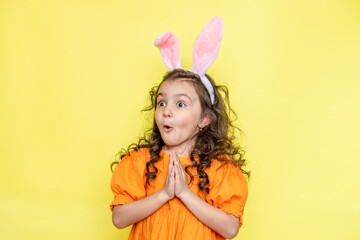 Obraz na płótnie Canvas Happy curly little girl wears bunny ears, loves the celebration and mystery of the holiday, dressed in orange bright dress, isolated on yellow background. Seasonal holiday concept 