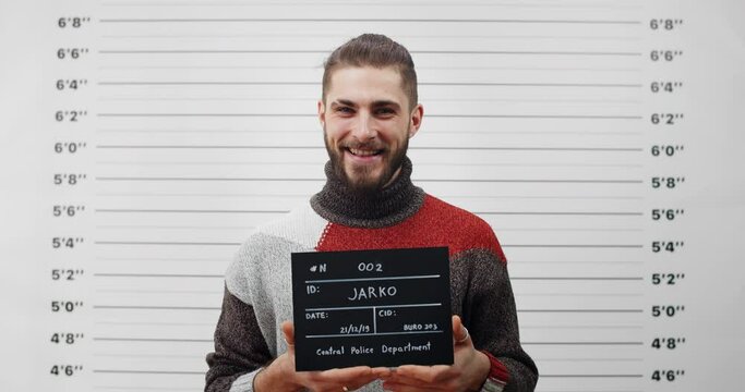 Portrait of handsome male criminal with beard holding sign for photo in police department. Crop view of young man posing and smiling while looking to camera.Concept of crime.
