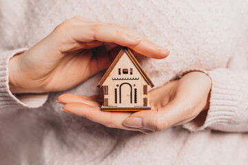 Woman holding a wooden house with her hands with the sun on a light pink background. Sweet home