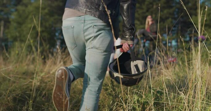 Close up of hipster man holding hemlet and going towards group of friend that are having fun in morning sun beside off road car