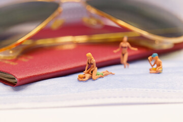 Figures sitting on a disposable surgical mask with sunglasses and a German passport in the background, waiting for summer vacation to travel again.
