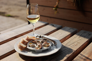 Glass with wine and dish with the snail shells, served on a white classic plate, with various sauces, pesto, truffle, cheese, pieces of French baguette, fork for a shell, wooden table in outdoor cafe