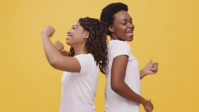 Two happy young african american women dancing, having fun together, standing back to back, orange background,