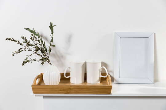 Dry branch of eucalyptus in a white figured earthenware vase. A two tall cups for tea in the tray on the table. Scandinavian style. White photo frame with blank space