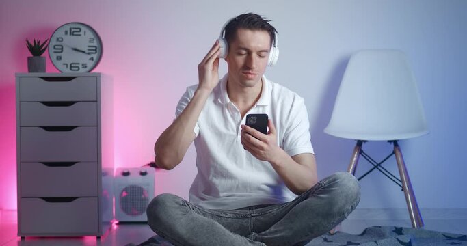Portrait of young stylish man enjoying listening to music in white headphones on smart phone and waving his head to the rythm while sitting on the floor at home.