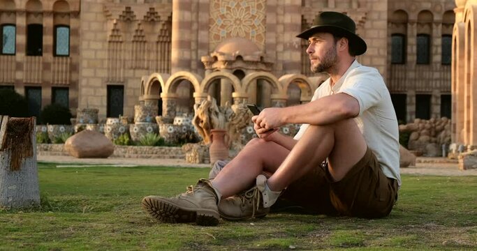 handsome tourist in boots and a cowboy hat, sits on the green grass, writes SMS,a broad-shouldered hiker in a shirt is resting after a long hike.concept archeology, geology, tourism, travel