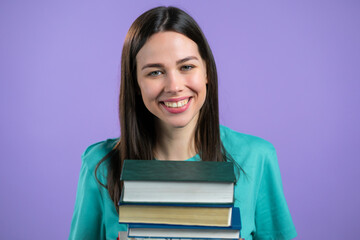 Student holds stack of university books from library on violet background in studio. Woman smiles, she is happy to graduate.