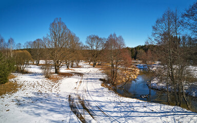 Early spring landscape with melting snow. Dirt rural road, wood, ice and snow panorama.