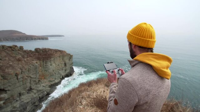 A man standing on the cliff edge is making video with drone of beautiful marine landscape and sea waves crashing with foam. 