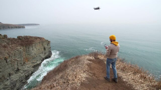 A man is standing on the picturesque cliff edge with drone panel control landing the copter on his hand. Filming beautiful marine landscape