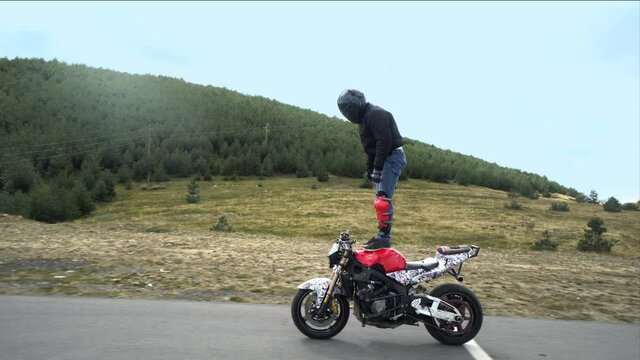 Stunt biker standing still on the moving motorcycle in the empty mountain road