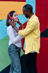 young multiethnic couple in love laughing while listening to music with red headphones and a red phone against a colorful background dressed in colorful clothes.