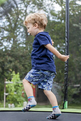 Fototapeta premium A cute little boy with blonde hair and blue eyes wearing a blue T-shirt and short shorts jumping on a trampoline. In the background, green leaves of trees and other vegetation.