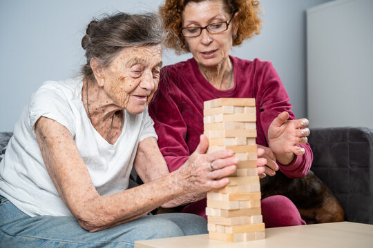 Elder woman and her adult daughter together with dachshund dog spend time together at home playing board game collecting wooden blocks in tower. Jenga game. Theme is dementia and alzheimer's