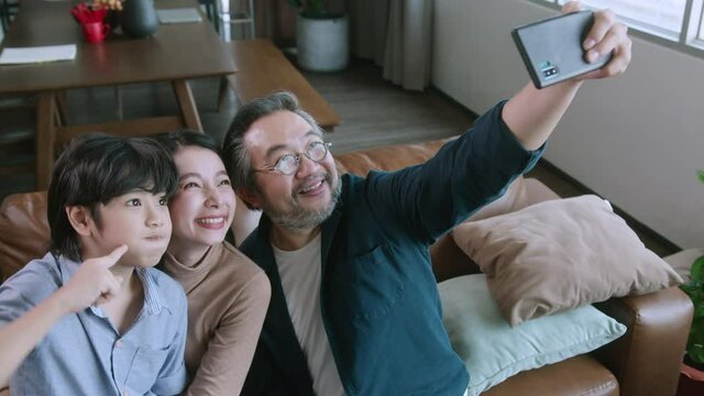 happiness joyful family love concept Asian family holiday activity dad holding smartphone taking a selfie photo together mom son and dad in the living room home interior background