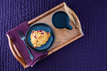 Breakfast in bed, homemade cranberry scone on black plate and wood tray on a purple bedspread
