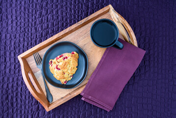 Breakfast in bed, homemade cranberry scone on black plate and wood tray on a purple bedspread
