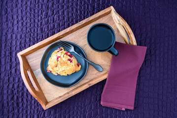 Breakfast in bed, homemade cranberry scone on black plate and wood tray on a purple bedspread
