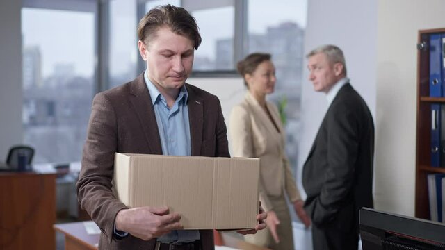 Portrait of sad fired young man standing in office with box as blurred colleagues talking at background. Stressed upset Caucasian employee leaving workplace. Employment and incompetence