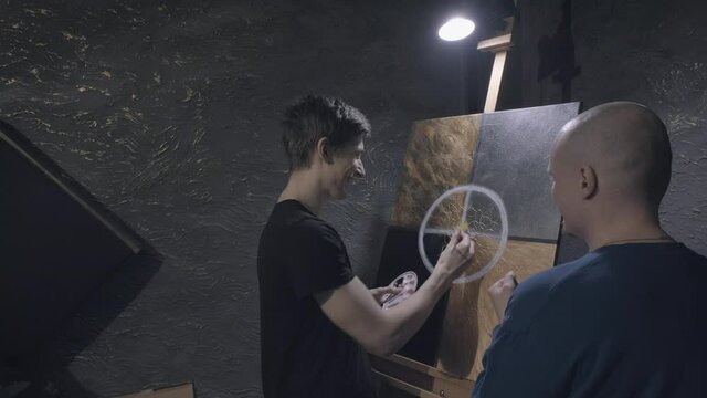 Two young, European artists discuss a beautiful, abstract picture. They enthusiastically communicate and laugh. The concept of teaching visual arts, constructive criticism and advice
