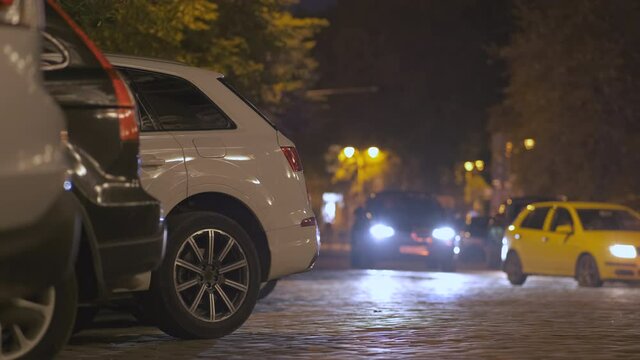 Close up of parked cars on roadside at night with blurred view of traffic lights of moving vehicles on city street.