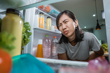 portrait of starving young asian woman hold her belly while open the fridge door looking for something to eat