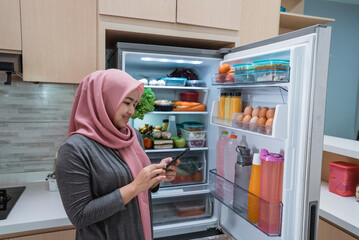 asian muslim young woman using her smartphone to buy groceries while open her fridge at home