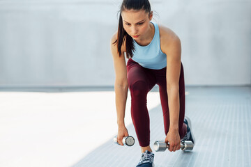 Horizontal image of an athelte young woman doing exercises with dumbells outdoors on a sunny day at grey background. Fit female squats with dumbbells outside.