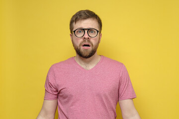 Frightened, shocked bearded man with glasses looks at the camera with large eyes and an open mouth. Yellow background.