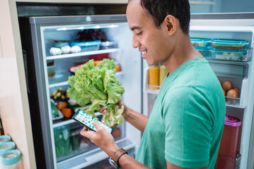 happy smiling man ordering online groceries shop. man standing in front of the fridge while holding his mobile phone