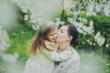 Fototapeta premium Happy mother's day. Little girl hugs her mother in the spring cherry garden. Portrait of happy mother and daughter among white flowers trees. Family values. Motherhood. Mom and child in blooming park.